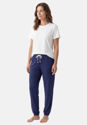 Kvinde stående barfodet iført en enkel hvid kortærmet t-shirt og mørkeblå joggerbukser med snøre mod en ensfarvet baggrund.
