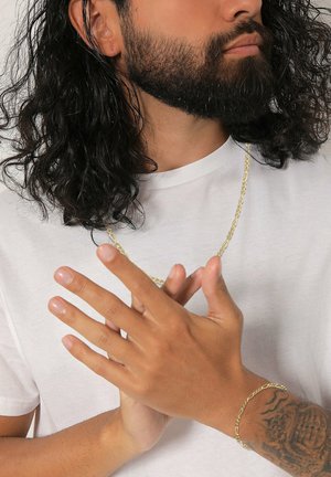Un collier en chaîne dorée repose sur un t-shirt blanc. Des mains jointes montrent un bracelet doré assorti. Les caractéristiques mises en avant comprennent des surfaces lisses et des motifs torsadés.