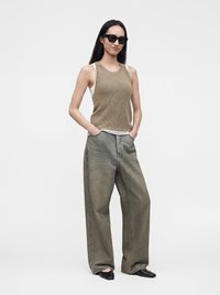 Femme portant des lunettes de soleil noires, un top en tricot beige sans manches par-dessus une chemise blanche, un pantalon vert délavé ample et des chaussures noires à bout carré, les mains dans les poches.