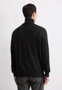 Mann in einem schwarzen Rollkragenpullover und grauen Hosen, der mit dem Rücken zur Kamera vor einer weißen Wand steht.