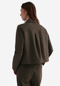 Braune cropped Jacke mit strukturierter Oberfläche, ausgestattet mit einem Reverskragen und einem gefältelten Rückendesign, kombiniert mit passenden Hosen.