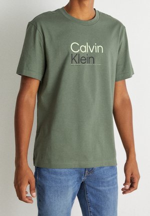 Person trägt ein olivgrünes Calvin Klein T-Shirt mit kurzen Ärmeln und blaue Jeans, steht vor einem schlichten hellen Hintergrund.