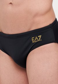 Črne plavalne hlače z gladko teksturo, s zlatim logotipom EA7 Emporio Armani na strani. Klasična zasnova z nizkim rezom.
