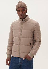 Beige gewatteerde jas met een hoge kraag, ritsluiting en zijzakken. Voorzien van ribgebreide manchetten en een gladde textuur. Gedragen over een blauwe spijkerbroek.