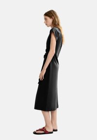 Zwart midi-jurk met korte mouwen, tailleband en een relaxte pasvorm. Stijlvolle rode sandalen met een gesp en een platte zool maken de outfit compleet.