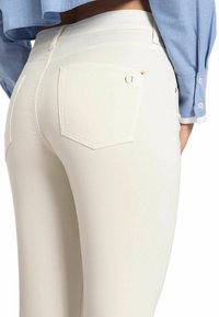 Witte jeans met een gladde textuur, vijf-pocket ontwerp en accenten van goudkleurig beslag, met een close-upweergave van de achterkant en stikseldetails.