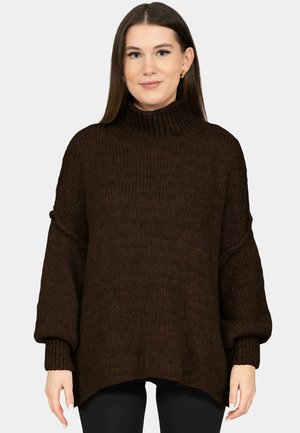 Kobieta ubrana w ciemnobrązowy, oversize'owy sweter z wysokim golfem i długimi bufkami na rękawach, w połączeniu z czarnymi spodniami.