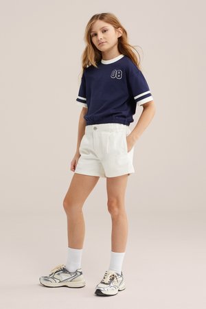 Mladé dievča stojí s jednou rukou v vrecku, oblečené v námornícky modrý top s krátkym rukávom s číslicou „08“, biele šortky, biele ponožky a sivé tenisky.