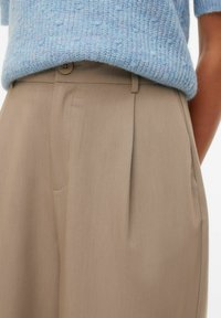 Femme portant un pantalon beige taille haute plissé et un pull en maille bleu clair rentré à l'intérieur.