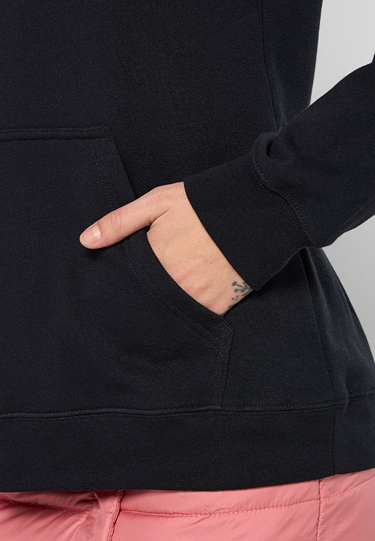 Hand mit kleinem Tattoo in der Tasche eines schwarzen Sweatshirts, das über rosa gesteppten Hosen getragen wird.