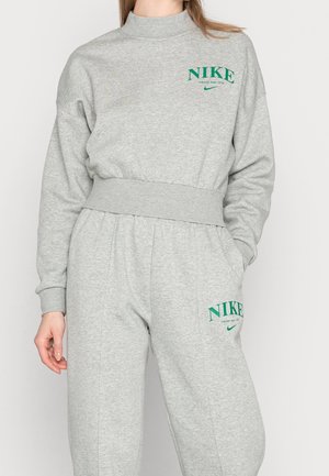 Kobieta ubrana w jasnoszary sweter Nike i pasujące spodnie dresowe z zielonymi logo Nike oraz napisami na klatce piersiowej i udzie.