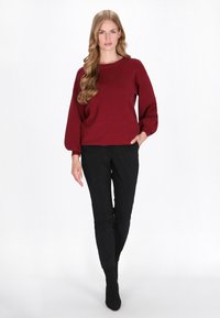Burgunderfarbener Strickpullover mit langen Ballonärmeln und rundem Halsausschnitt. Kombiniert mit schwarzer, eng anliegender Hose und knöchellangen schwarzen Socken.