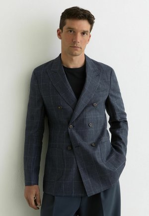 Mężczyzna w ciemnoniebieskiej dwurzędowej marynarce w kratę na czarnej koszuli, stojący na tle gładkiej, białej ściany.
