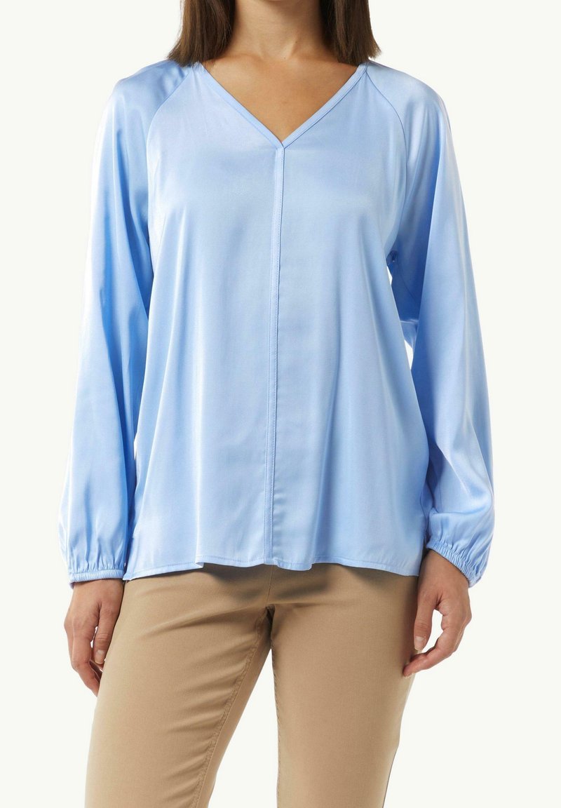 Femme portant un chemisier bleu clair à manches longues et col en V, associé à un pantalon beige, debout devant un fond clair uni.