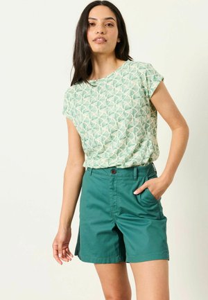 Frau mit langen dunklen Haaren, die ein kurzärmeliges Oberteil mit grünem Blattmuster und einfarbige türkisfarbene Shorts trägt, mit einer Hand in der Tasche.