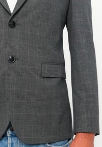 Hallgriseeritud ruuduline bleiser mustade nööpidega, seljas helesiniste teksade kohal, näidates esist pocket flap'i ja osa kandja käest.