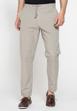 Uomo che indossa pantaloni beige casual con vita con coulisse e mocassini in pelle marrone, in piedi davanti a uno sfondo semplice.