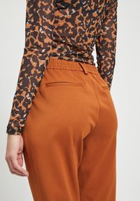 Persona que lleva pantalones elásticos marrones con cintura elástica y una blusa de manga larga con estampado de leopardo en negro y marrón, vista desde atrás a la altura de la cintura.