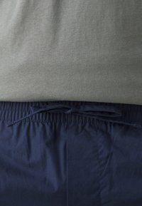 Zone de la taille d'une personne portant une chemise grise rentrée dans un pantalon bleu marine avec une taille à cordon noué.