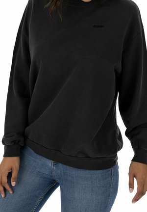 Personne portant un sweat-shirt noir uni à manches longues et un jean bleu, debout les bras détendus le long du corps.