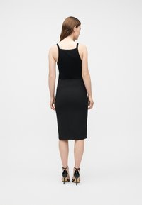 Γυναίκα που στέκεται με την πλάτη στραμμένη, φοράει μαύρο αμάνικο τοπ, εφαρμοστή μαύρη φούστα μήκους μέχρι το γόνατο και μαύρα πέδιλα με ψηλό τακούνι σε λευκό φόντο.