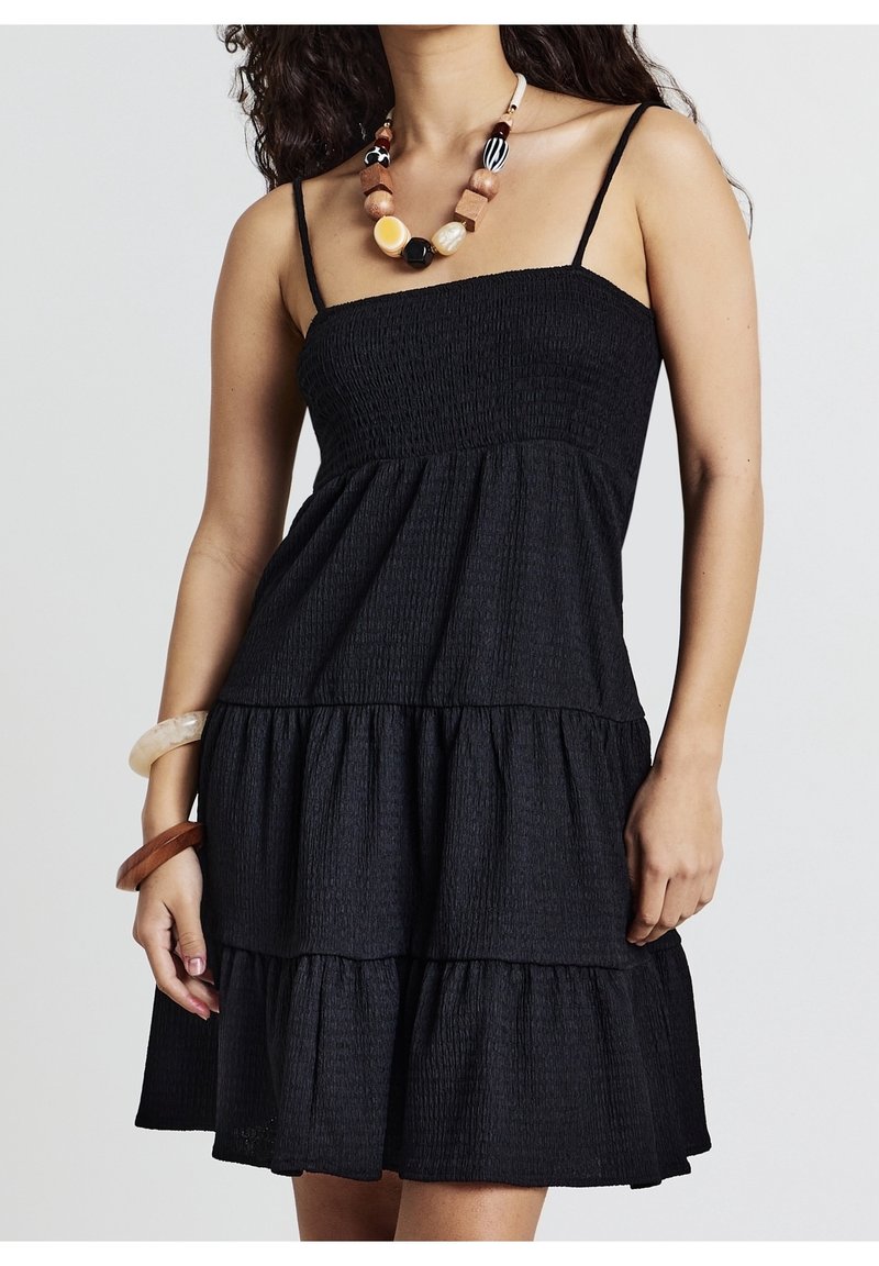 Robe noire à volants avec un corsage smocké et des bretelles fines. Texture douce avec un design fluide. Le mannequin porte un collier coloré en accessoire.