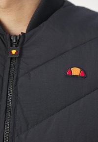 Μαύρο καπιτονέ μπουφάν με διαγώνια ραφή, που διαθέτει κλείσιμο με φερμουάρ και πολύχρωμο λογότυπο με κόκκινες, κίτρινες και πορτοκαλί λεπτομέρειες.