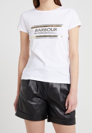 Kobieta w białym t-shircie z krótkim rękawem i logo Barbour International, w połączeniu z czarnymi skórzanymi szortami z podwiniętymi nogawkami, stojąca na jednolitym tle.