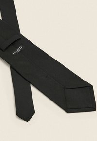 Musta solmio, joka on valmistettu teksturoidusta kankaasta ja jossa on terävä pää, sisältää logon, jossa lukee "HACKETT" pienellä lenkillä. Klassinen muotoilu.