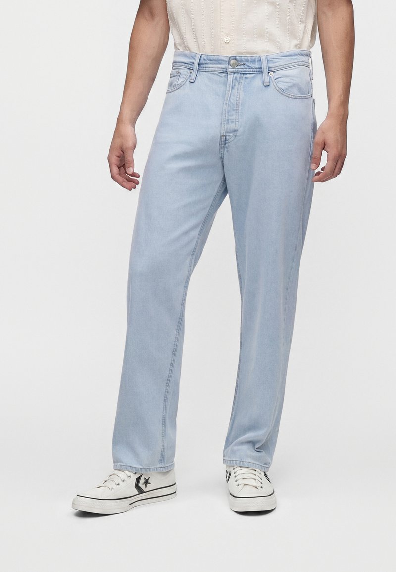 Mann trägt hellblaue Straight-Leg-Jeans mit Fronttaschen und weiße Sneakers mit schwarzem Stern-Detail, steht auf weißem Hintergrund.