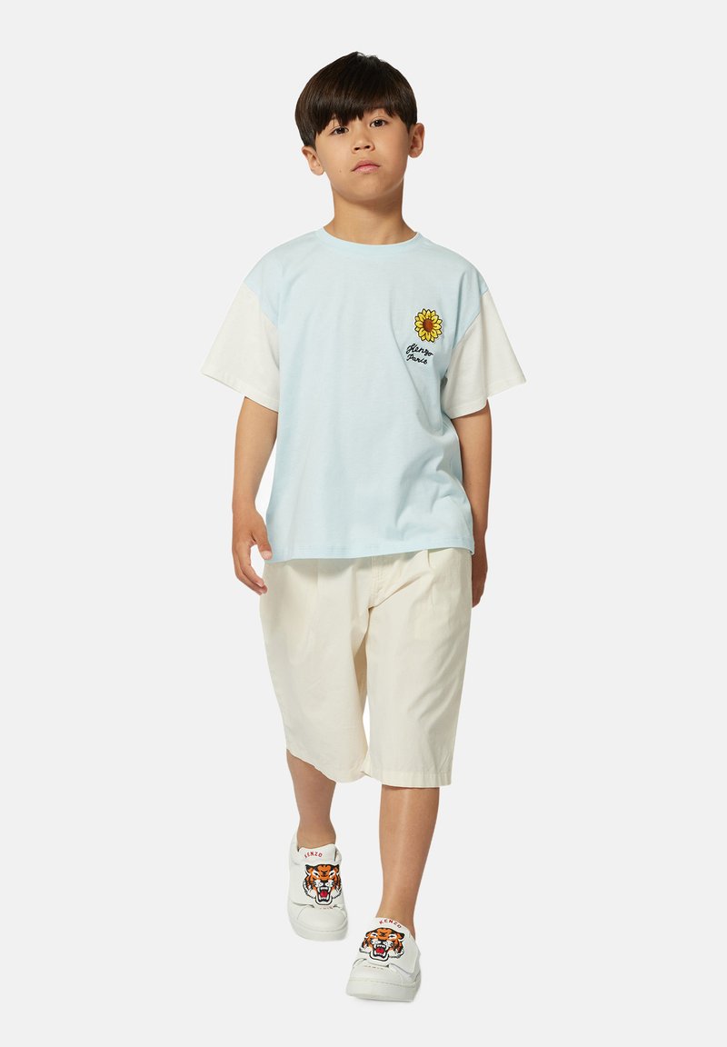 T-shirt bleu clair avec des manches blanches, broderie de tournesol, associé à un short crème et des baskets blanches avec un design de tigre.