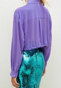 Blusa trasparente viola con maniche lunghe e colletto alto, abbinata a una gonna di paillettes verdi scintillanti. La blusa presenta un orlo arricciato e dettagli con bottoni.