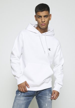 Jaunas vyras dėvi baltą džemperį su gobtuvu su "cK" logotipu ir dėvėtus mėlynus džinsus, stovintis priešais paprastą šviesų foną.