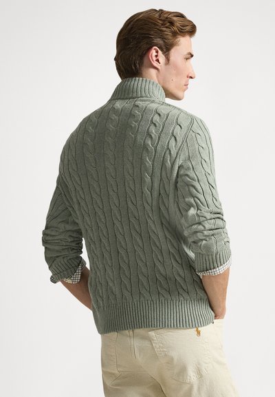 Light green cable-knit sweater with a high collar and ribbed hem. Features rolled sleeves and a checkered shirt peeking out.