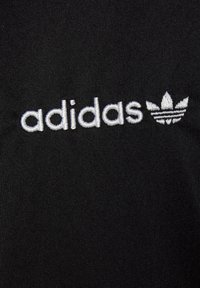 Fekete szövet, textúrált felülettel, rajta egy fehér hímzett "adidas" logóval és alatta egy háromlevelű emblémával.