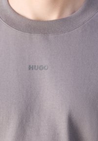 Gros plan d'une personne portant un t-shirt gris col rond avec le mot "HUGO" imprimé en petites lettres foncées sur la poitrine.