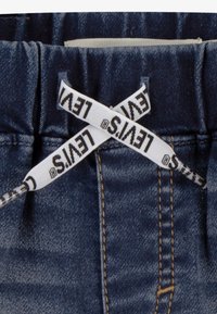 Ceinture en denim bleu foncé avec un élastique, ornée d'un ruban blanc Levi's noué en nœud. Couture jaune le long des bords.