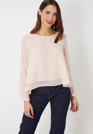 Mujer con cabello castaño, vestida con blusa de manga larga en capas de color rosa claro y vaqueros azul oscuro, sonriendo con lápiz labial rojo, de pie frente a un fondo blanco.