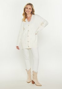 Weißer Strickcardigan mit Holzknöpfen, V-Ausschnitt und langen Ärmeln, kombiniert mit weißen Hosen und beigen, im Western-Stil gehaltenen Stiefeletten.