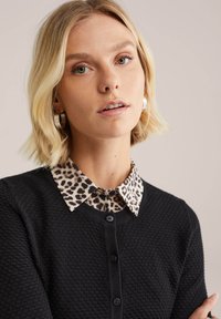 Plava žena s kratkom kosom nosi srebrne naušnice na prsten, ovratnik s leopard uzorkom i crni strukturirani kardigan na dugmad, gleda unaprijed.
