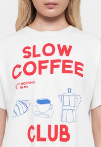 Balta T-kreklu ar sarkanu tekstu "SLOW COFFEE CLUB" un maziem ziliem attēliem ar kruasānu, kafijas krūzi uz šķīvīša un kafijas katlu.
