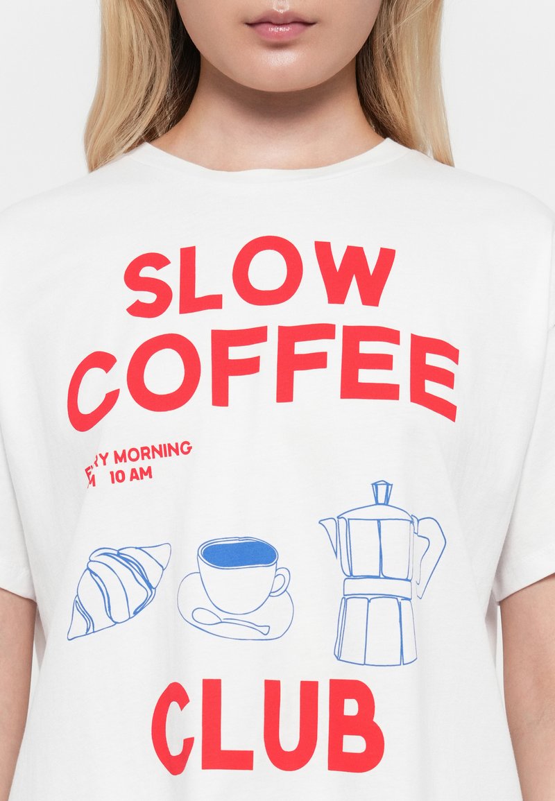 T-shirt bianca con testo rosso "SLOW COFFEE CLUB" e piccole illustrazioni blu di un cornetto, una tazza di caffè su un piattino e una caffettiera.