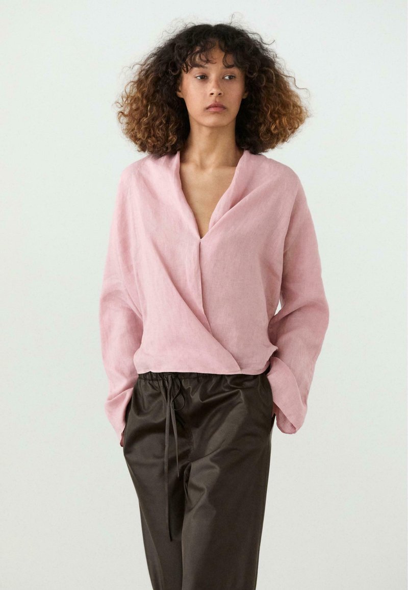 Vrouw met krullend haar die een losse roze wikkelblouse en donkere broek met koord draagt, staand tegen een eenvoudige lichte achtergrond.