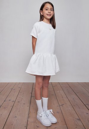 Ragazza giovane in piedi su pavimento in legno, indossa un vestito bianco corto, calzini bianchi e scarpe da ginnastica bianche, sorride con le mani dietro la schiena.