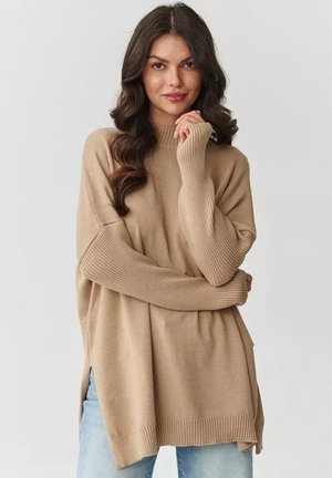Beżowy, oversize sweter z wysokim ściągaczem, ściągaczami na rękawach i bocznymi rozcięciami. Miękka tekstura, luźny fason, w połączeniu z jasnymi dżinsami.
