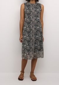 Robe sans manches mi-longue avec des rayures zèbre abstraites noires et beiges, portée avec des sandales à brides croisées couleur tan sur un fond clair.