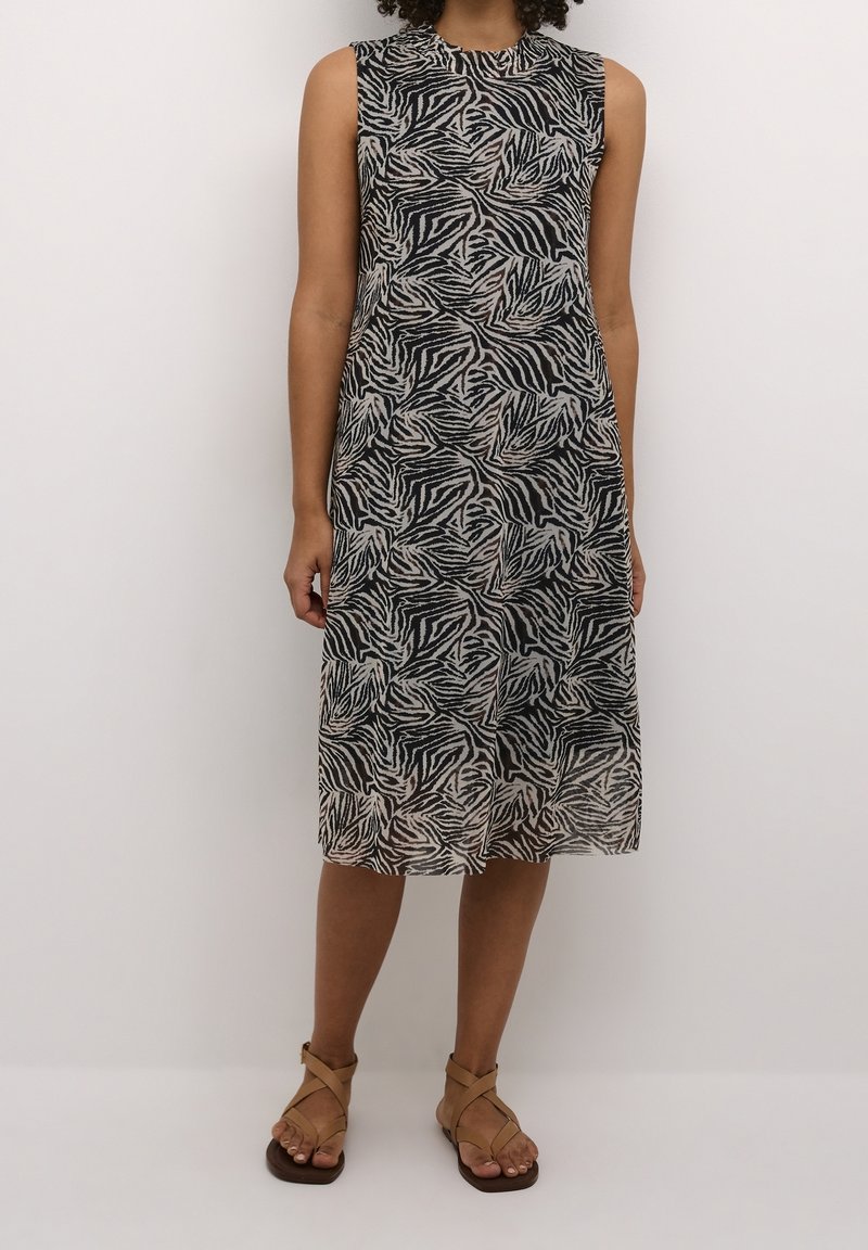 Robe sans manches mi-longue avec des rayures zèbre abstraites noires et beiges, portée avec des sandales à brides croisées couleur tan sur un fond clair.