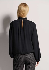 Schwarze Bluse mit Rüschenausschnitt und Schlüsselbeinausschnitt auf der Rückseite. Glattes Material, lange Ärmel und geraffte Manschettenakzente. Kombiniert mit strukturierten Hosen.