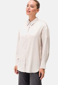 Blusa a maniche lunghe rosa chiaro con chiusura a bottoni frontale, colletto classico e tessuto morbido e liscio. Presenta una vestibilità rilassata e una leggera lucentezza.