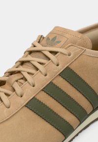 Μπεζ δερμάτινο αθλητικό παπούτσι με τρεις σκοτεινές πράσινες ρίγες, στρογγυλή μύτη, υφή στην επιφάνεια και επίπεδα κορδόνια με ανάγλυφο λογότυπο στη γλώσσα.